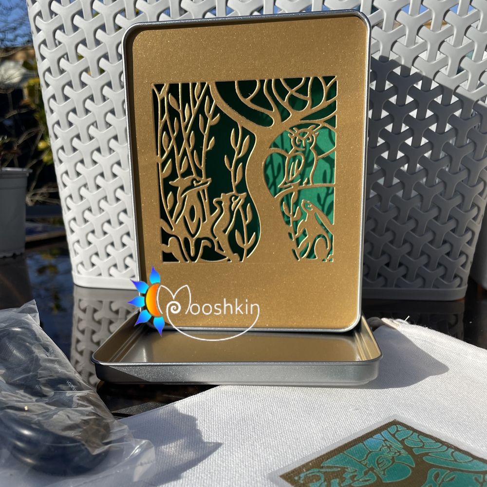 A Mooshkin Lantin featuring animals and trees die-cut into gold card, overlaid on green card, in a light-up scene inside a tin.