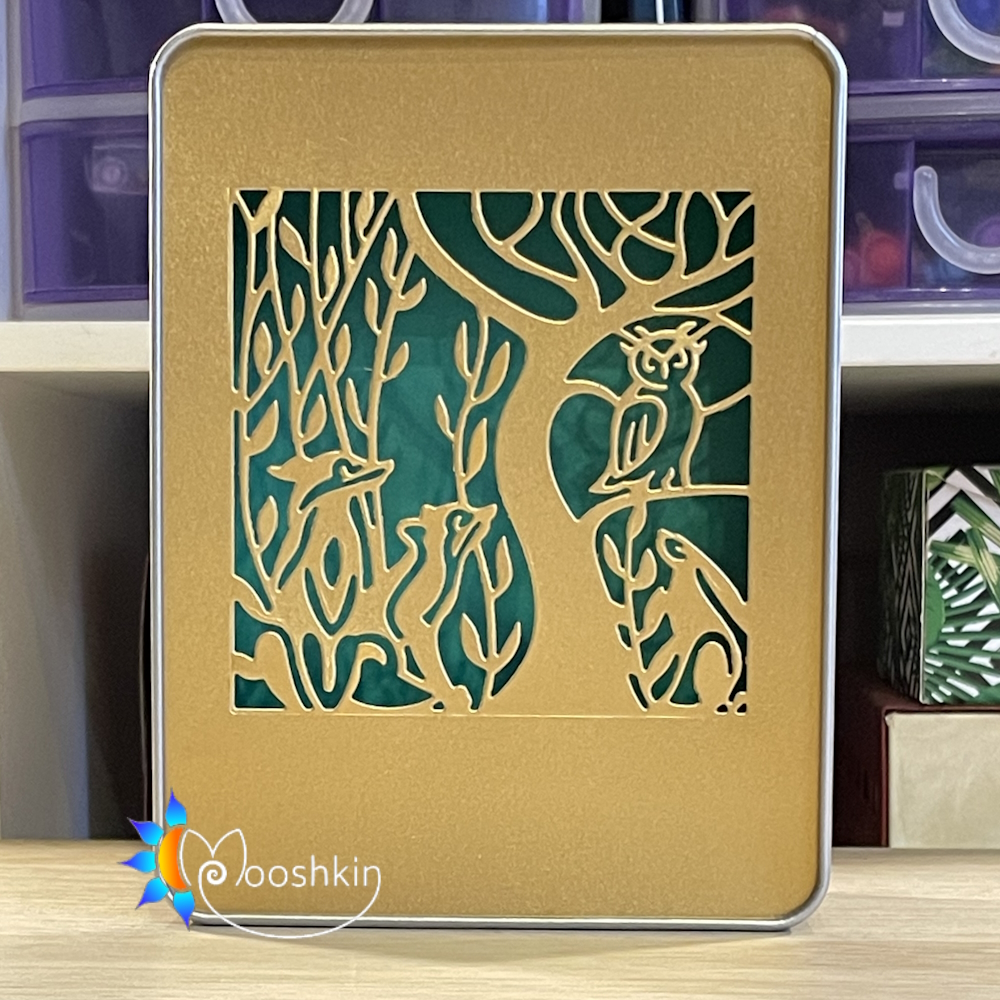 A Mooshkin Lantin featuring animals and trees die-cut into gold card, overlaid on green card, in a light-up scene inside a tin.